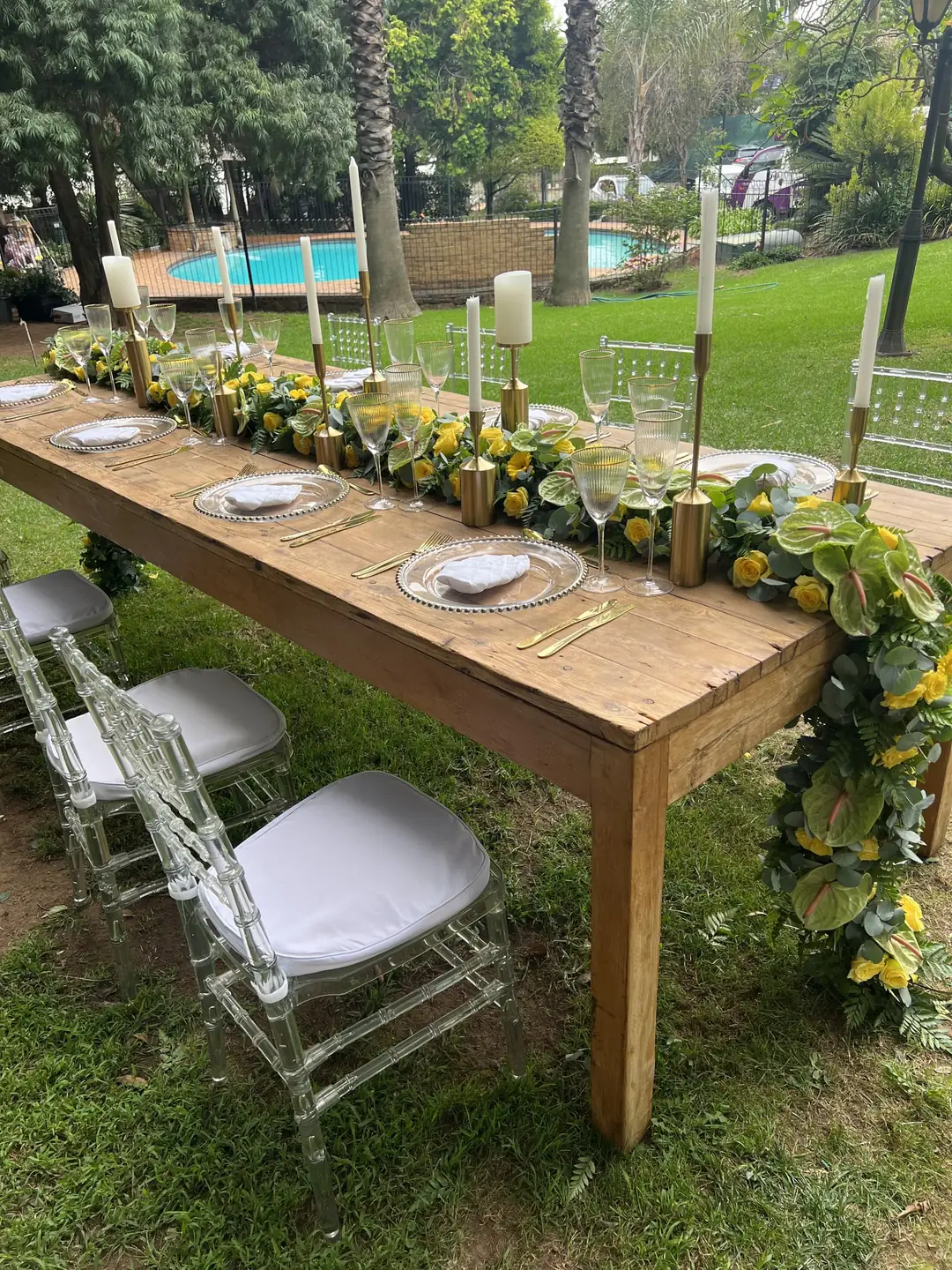 Rustic timber table with a cascading garland of yellow roses, lime anthuriums and eucalyptus between gold candlesticks, beside a garden pool