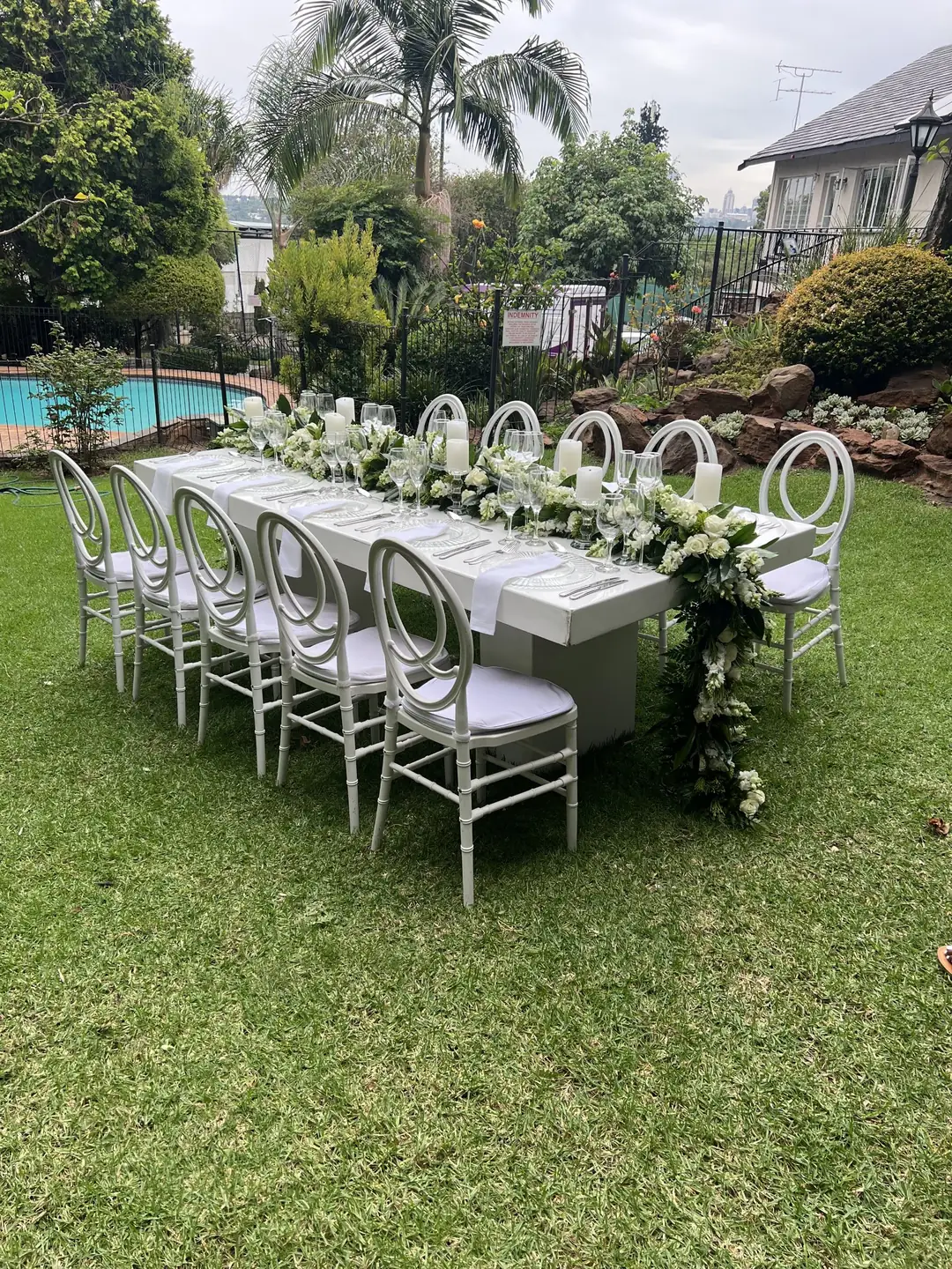 Long white table and white ring-back chairs, with a white rose and snapdragon garland spilling from the table end, beside a garden pool