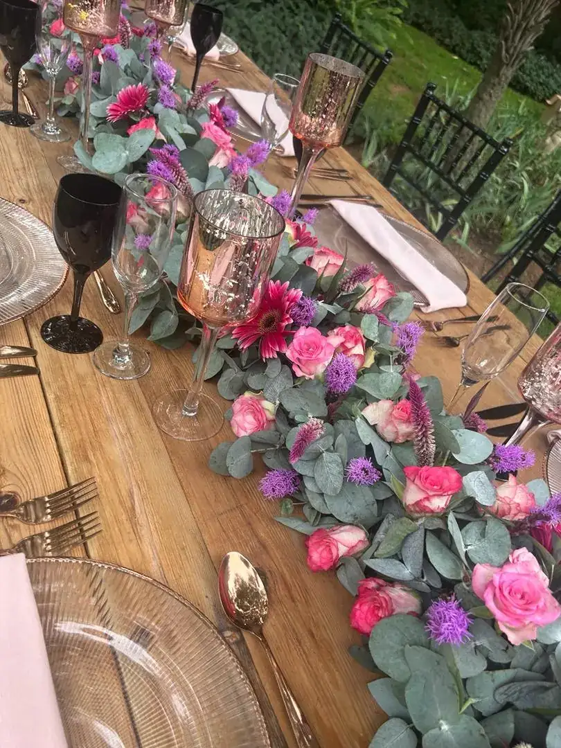 Table garland of pink roses, gerberas and purple blooms on eucalyptus, with mercury-glass goblets and black stemware on a timber table