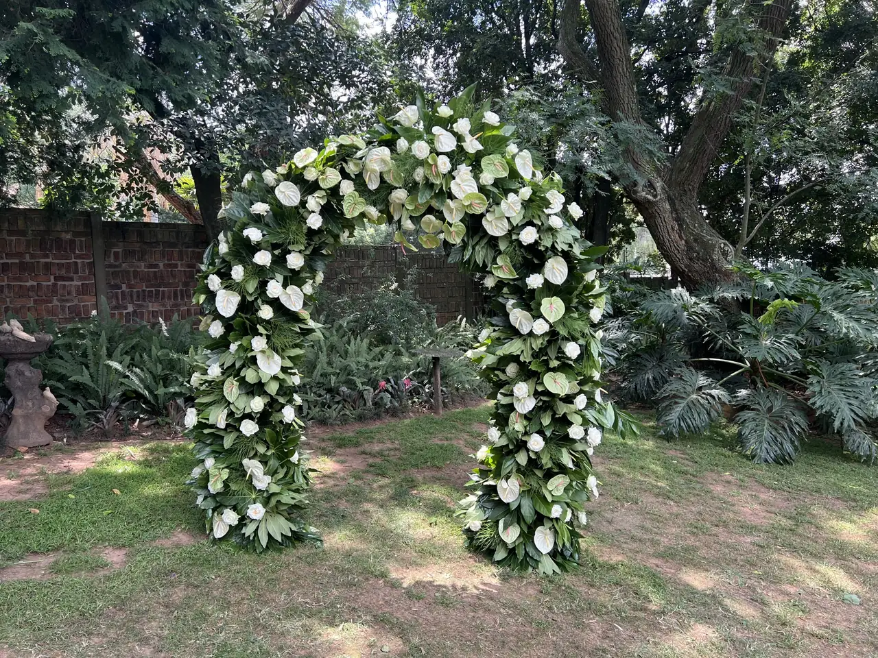 Tall floral arch covered in white roses, white and green anthuriums and tropical foliage, standing on a garden lawn