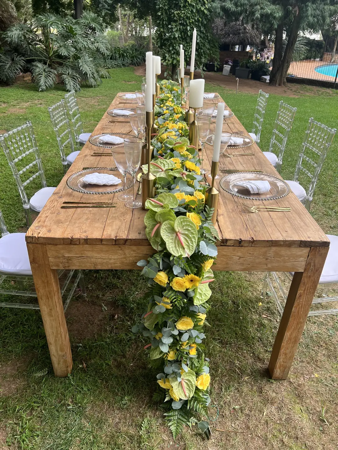 Rustic timber table seen along its length, with a yellow rose and lime anthurium garland between white tapered candles in gold holders