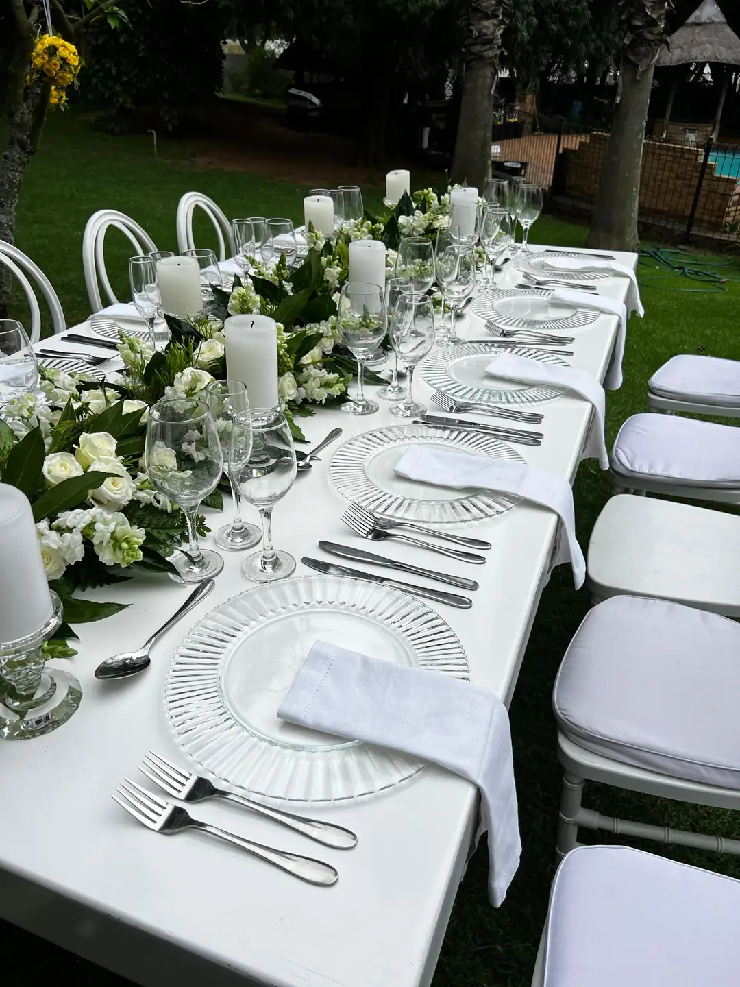 Place setting on a white table with a ribbed glass charger, linen napkin and silver cutlery, beside a white rose and snapdragon centrepiece