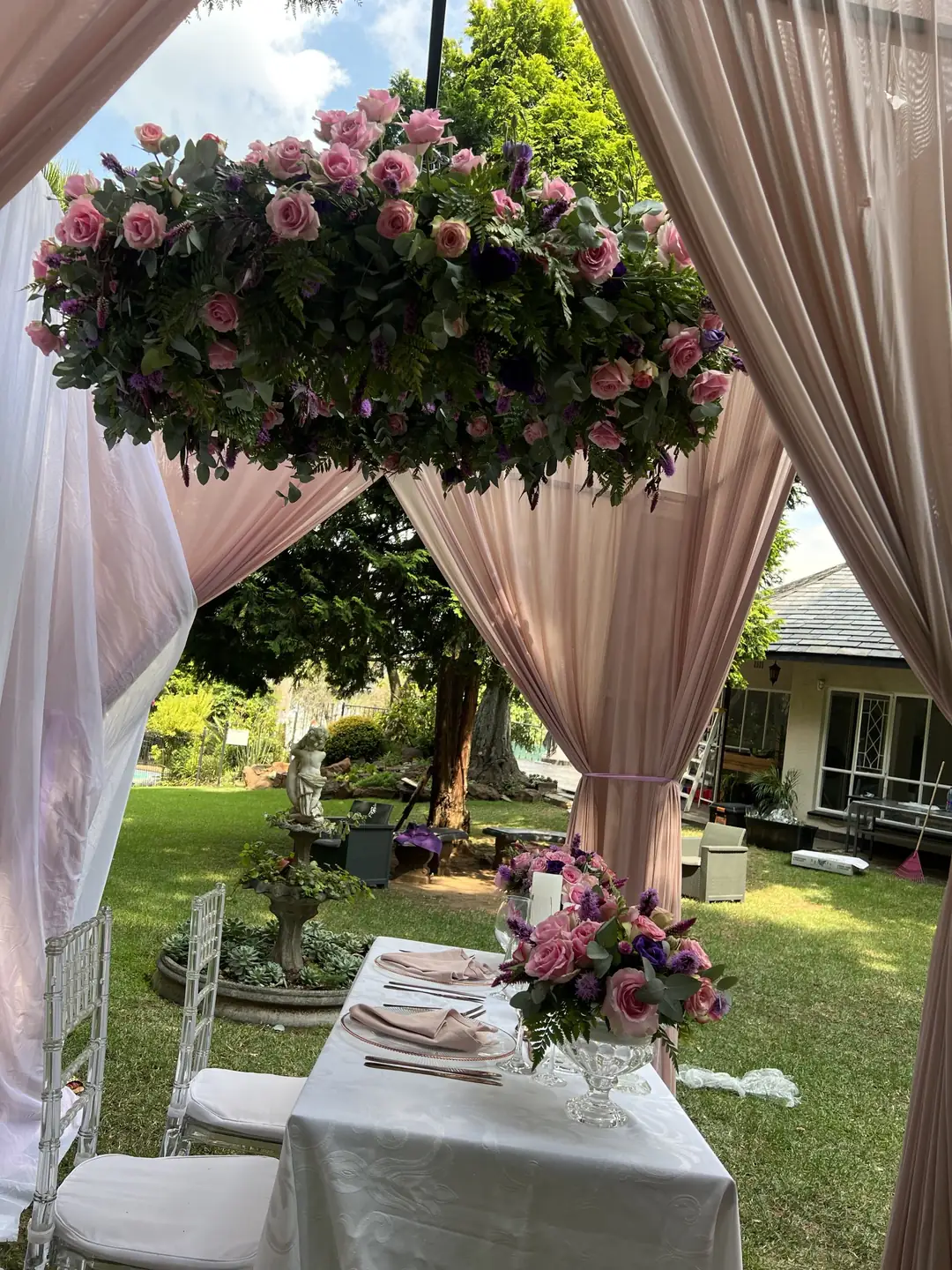 Hanging floral installation of pink roses and purple blooms above a blush-draped pavilion, with a small table set beneath