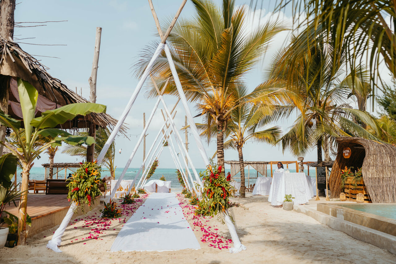 Beach wedding ceremony aisle styled with a draped arch and floral petals on sand, framed by palm trees