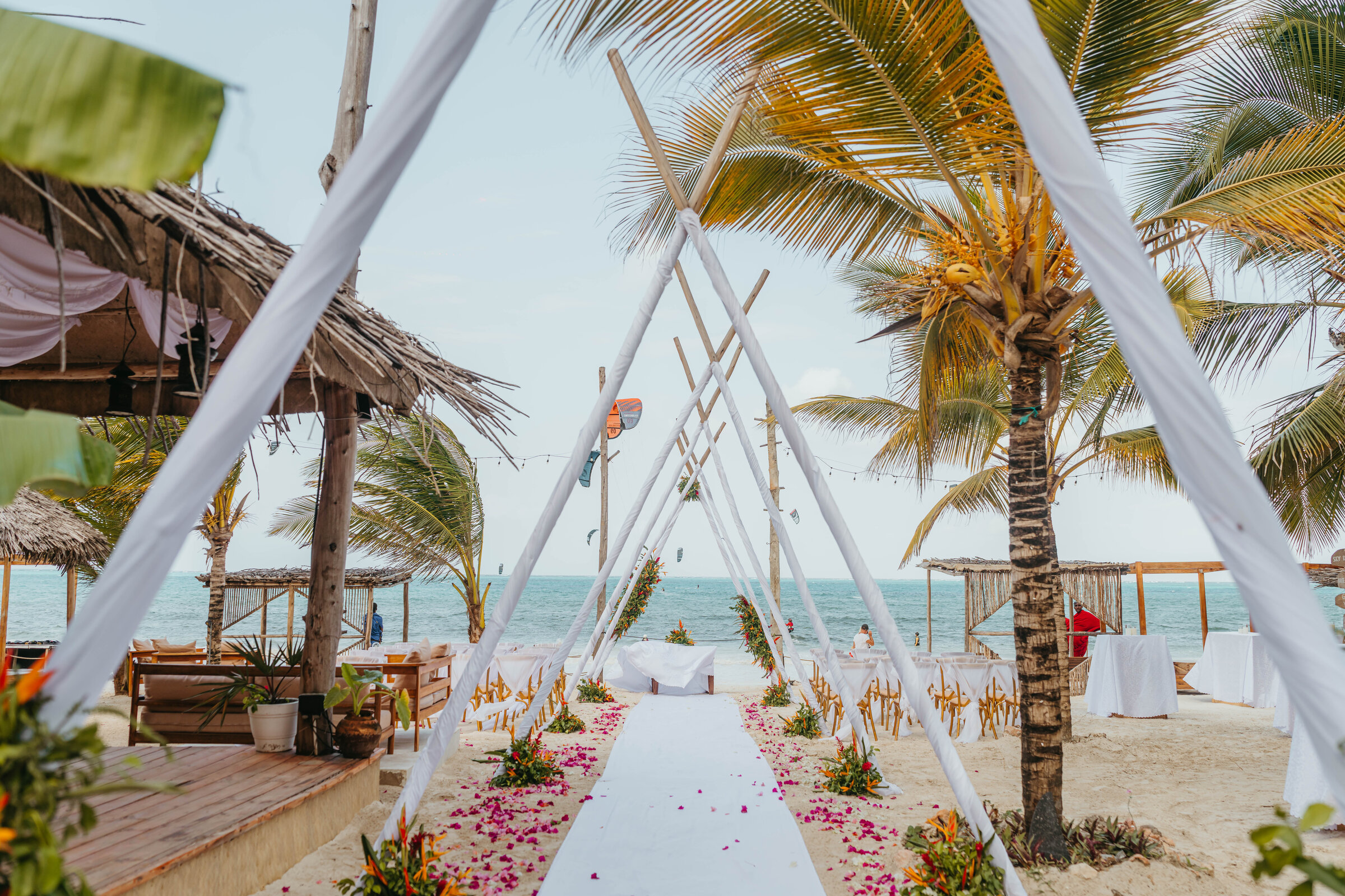 Draped teepee ceremony arch on a beach framed by palm fronds, with a petal-lined aisle to the ocean