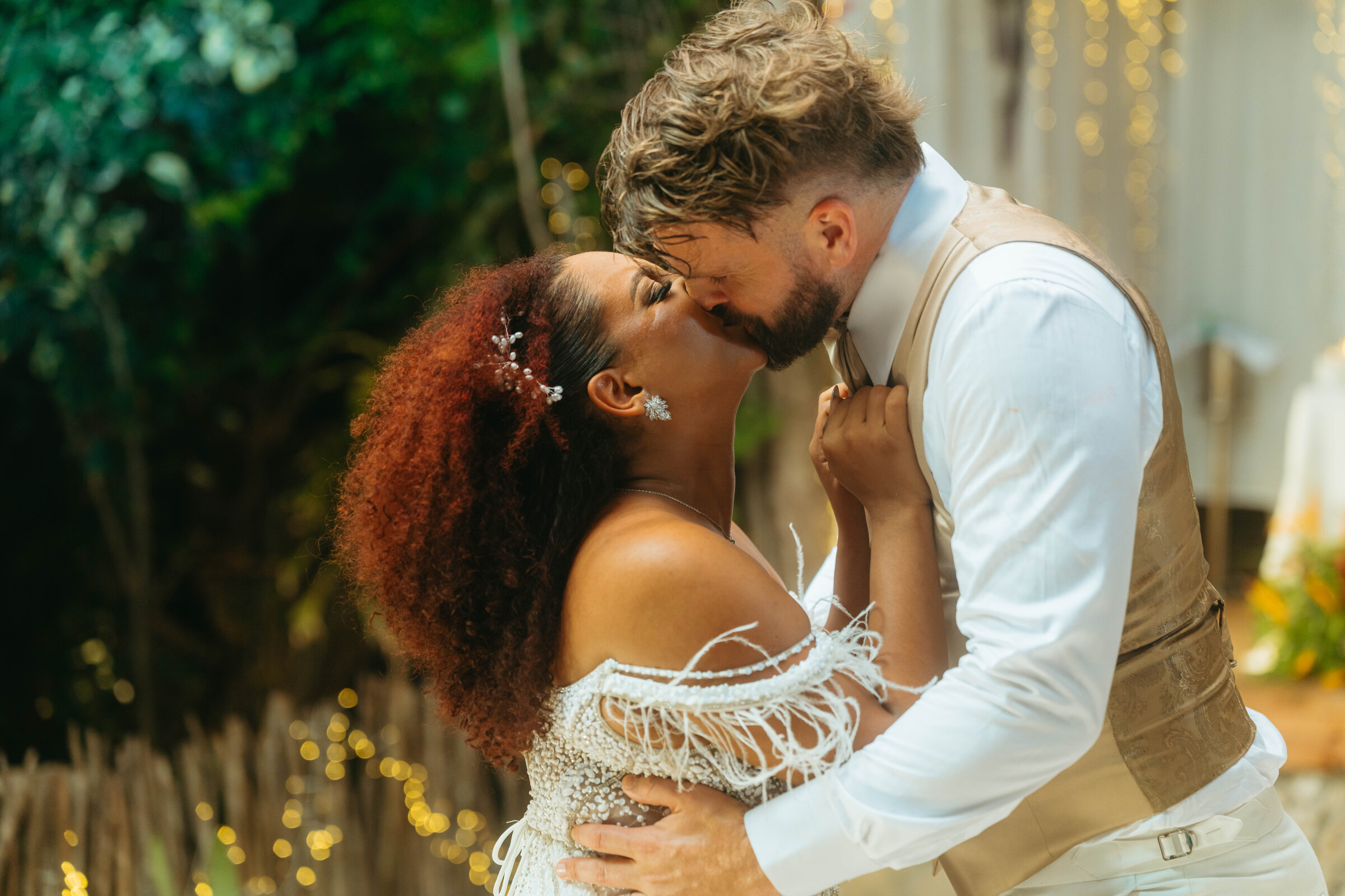 Bride and groom kissing at night surrounded by string lights and greenery