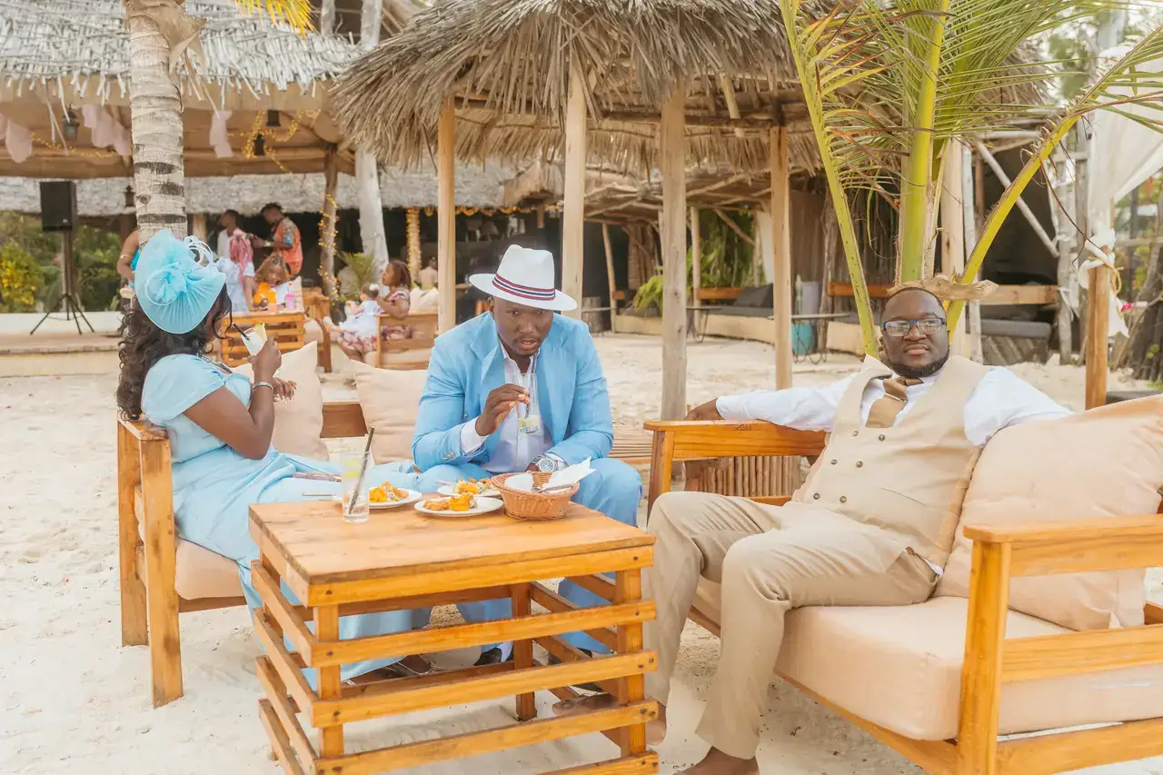 Guests relaxing on timber lounge seating under thatched shelters at a beach reception